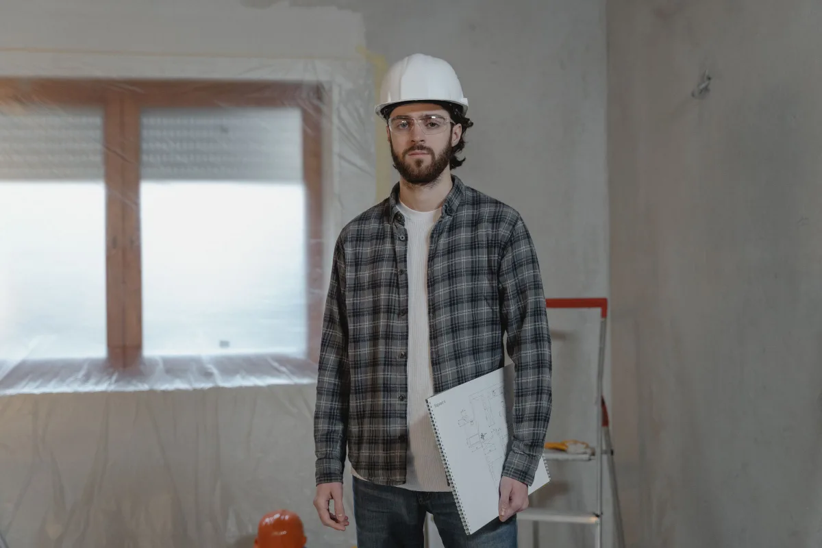 Drywall professional in a hard hat and safety glasses holding plans in a room masked off before work starts